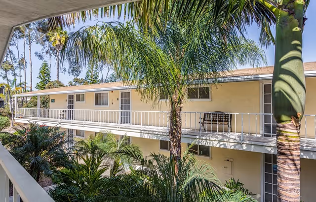 View of a two-story senior living facility building with exterior walkways and doors to individual units. The building is surrounded by lush green palm trees and other tropical plants under a clear blue sky.