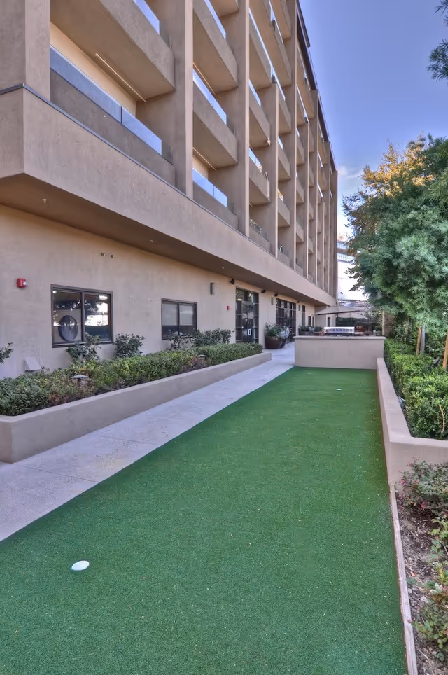 Outdoor area of a senior living facility with a long artificial grass putting green bordered by low concrete walls and shrubbery. The beige multi-story building with balconies is on the left side, and trees are on the right side under a clear blue sky.