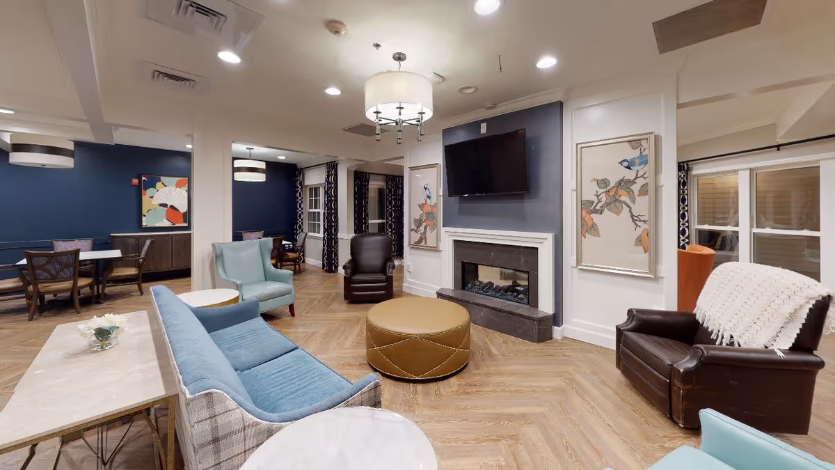 A cozy senior living facility common area with a blue sofa, two light blue armchairs, a brown leather armchair with a white knitted throw, a round tan ottoman, and a fireplace with a mounted flat-screen TV above it. The walls are decorated with framed artwork featuring birds and branches. In the background, there are dining tables and chairs against dark blue walls with patterned curtains on the windows.