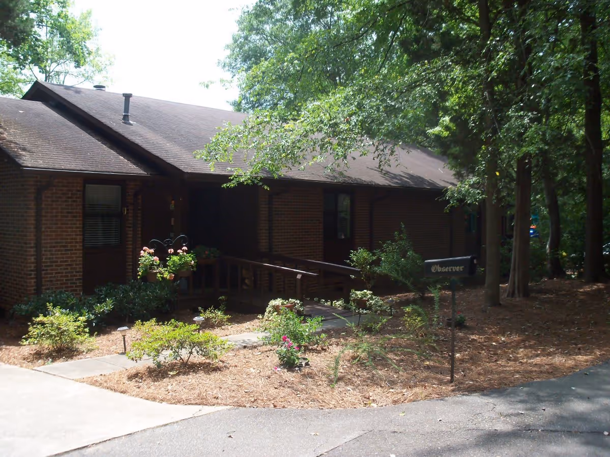 Exterior view of a single-story brick building surrounded by trees and landscaped garden beds with shrubs and flowers. A paved walkway leads to a ramp entrance with flower pots hanging on the railing. A small sign labeled 'Observer' is visible near the walkway.