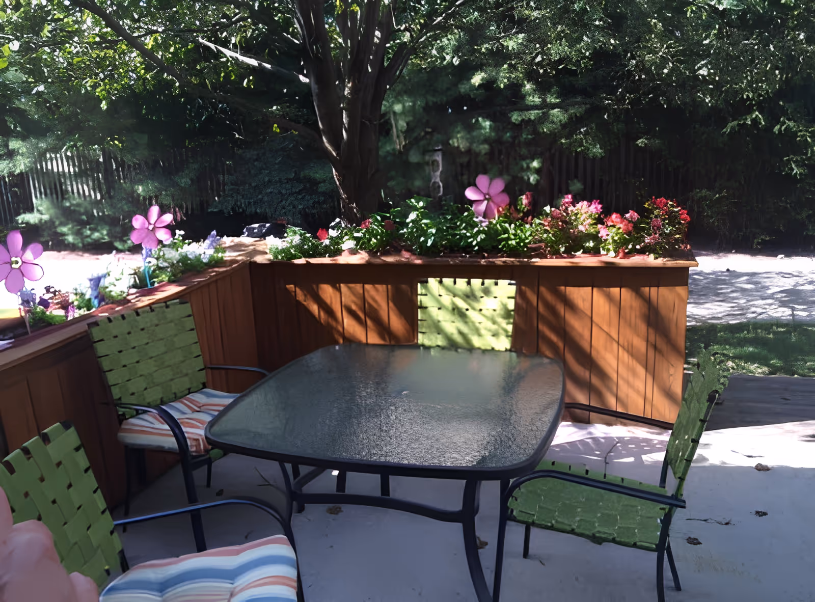 Outdoor patio with a glass-top table and green woven chairs beside wooden planter boxes full of flowering plants under trees.