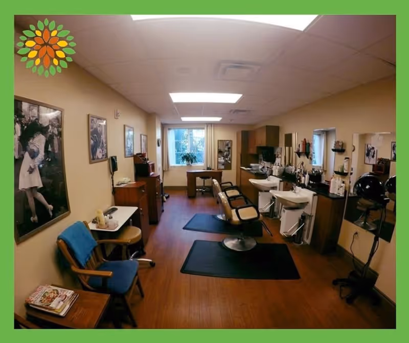 Interior view of a salon area in a senior living facility with two salon chairs in front of sinks, mirrors on the wall, hair dryers, and a small seating area with a chair and table. The room has wooden flooring, beige walls, and framed black and white photos on the walls. A window at the far end lets in natural light.