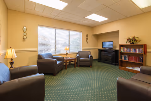 Well-lit sitting area with leather armchairs, a TV and bookshelf beside a large window.
