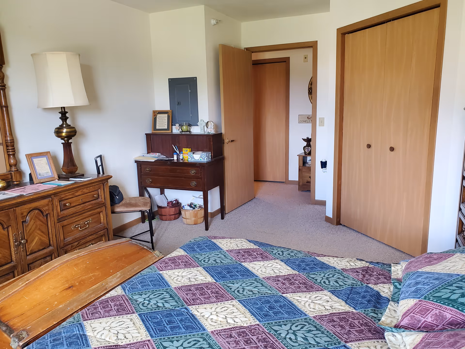 A bedroom with a bed covered in a colorful patterned quilt in the foreground. To the left, there is a wooden dresser with a lamp and framed pictures on top. A small wooden desk with drawers and various items, including a tissue box, is against the far wall. A chair is positioned next to the desk. The room has beige carpet and light-colored walls. There is a door open to a hallway and a closed double-door closet with wooden doors.