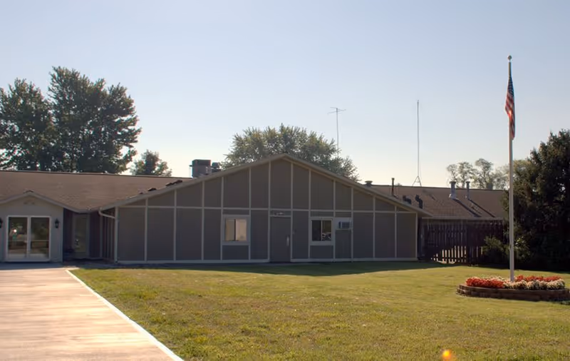 Front exterior of a single-story nursing facility with a lawn, flagpole, and paved walkway.