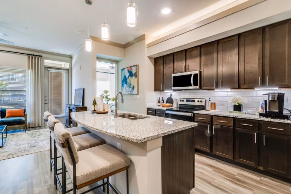 Modern kitchen with dark wood cabinets, granite countertops, and stainless steel appliances including a microwave and stove. A kitchen island with a sink and three cushioned bar stools is in the foreground. Pendant lights hang above the island. In the background, there is a living area with a blue chair, orange pillow, and large windows with curtains.