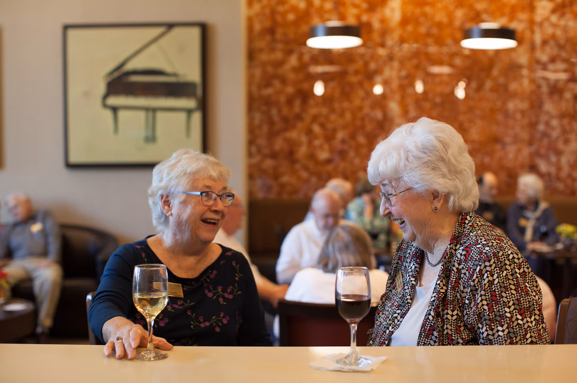 Two elderly women sitting at a table in a social setting, smiling and enjoying conversation with glasses of wine in front of them. Other seniors are seated and interacting in the background, with warm lighting and a framed picture of a piano on the wall.