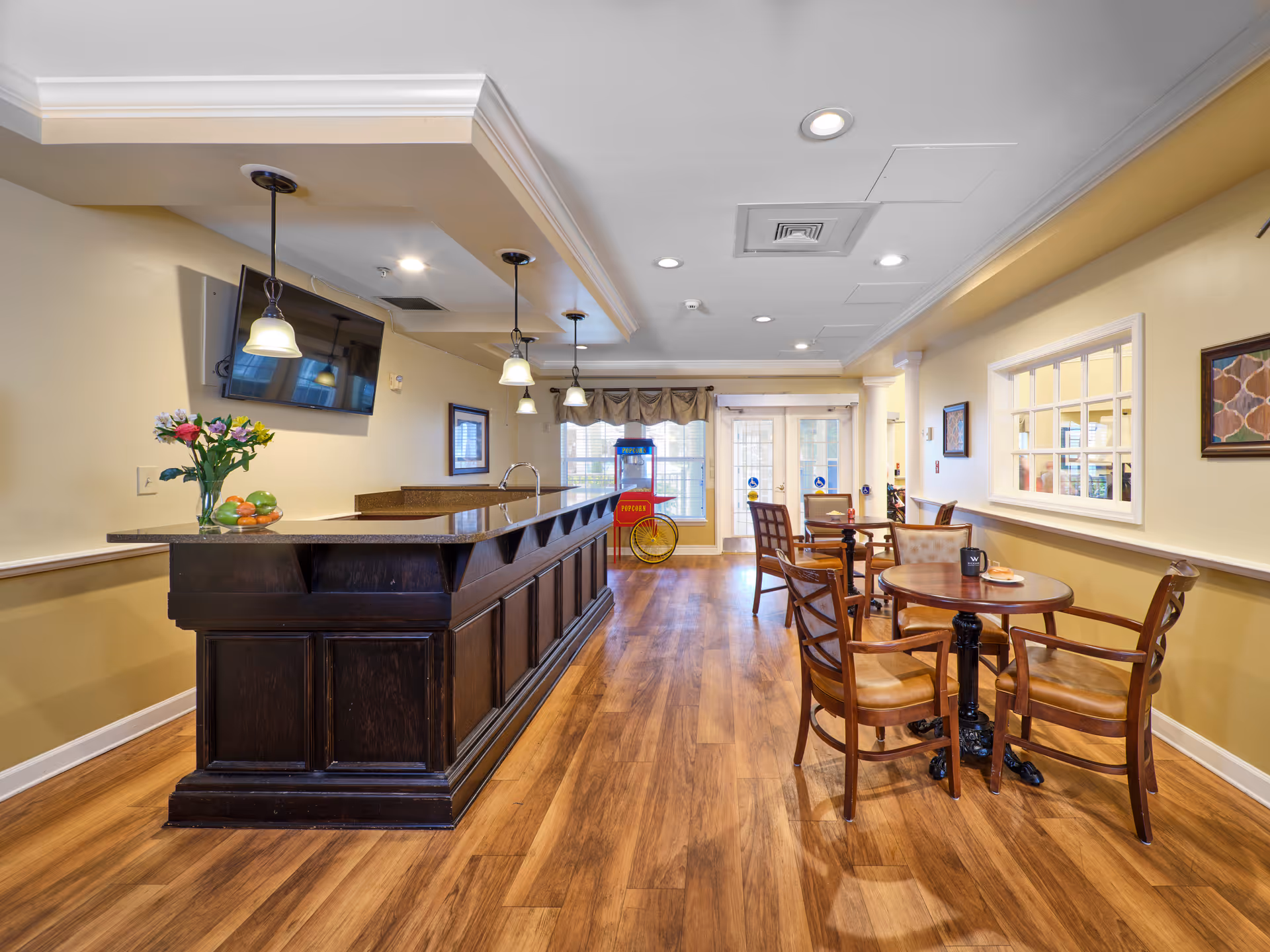 Interior view of a common area in Colliers Assisted Living at Port Orange featuring a dark wooden bar counter with pendant lights hanging above it, a wall-mounted TV, a vase with flowers and fruit on the counter, wooden flooring, and several round tables with chairs arranged for seating. In the background, there is a popcorn machine near glass doors leading outside.
