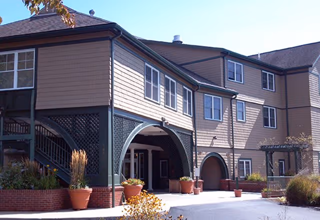 Exterior view of a multi-story senior living facility building with beige siding and green trim. The entrance features large green arches and a covered walkway. There are several large potted plants and some landscaping with bushes and ornamental grasses near the entrance.