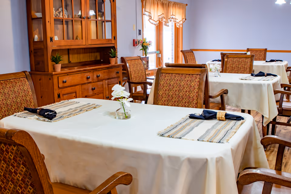 A dining room in a senior living facility with tables covered in white tablecloths, each set with placemats, napkins, and small flower vases. Wooden chairs with patterned upholstery surround the tables. A wooden cabinet with glass doors and decorative items is visible against the wall, and a window with curtains lets in natural light.