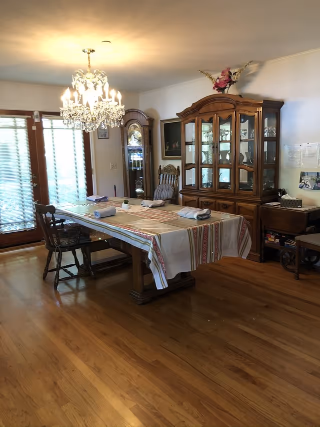 A dining room with a wooden table covered by a striped tablecloth and four rolled napkins placed on it. There are wooden chairs around the table, a large wooden china cabinet with glass doors displaying glassware, a grandfather clock, a chandelier hanging from the ceiling, and glass doors with blinds letting in natural light.