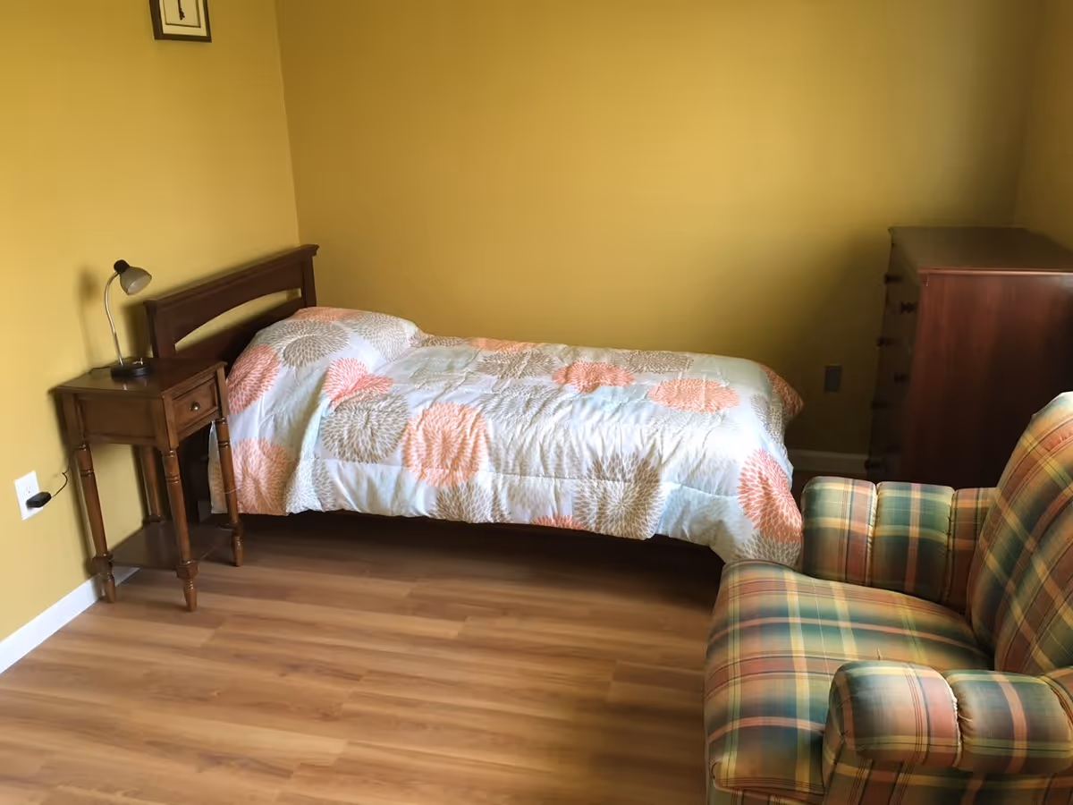 A small bedroom with a single bed covered with a floral patterned quilt, a wooden nightstand with a lamp, a wooden chest of drawers, and a plaid upholstered armchair. The walls are painted yellow and the floor is wood.