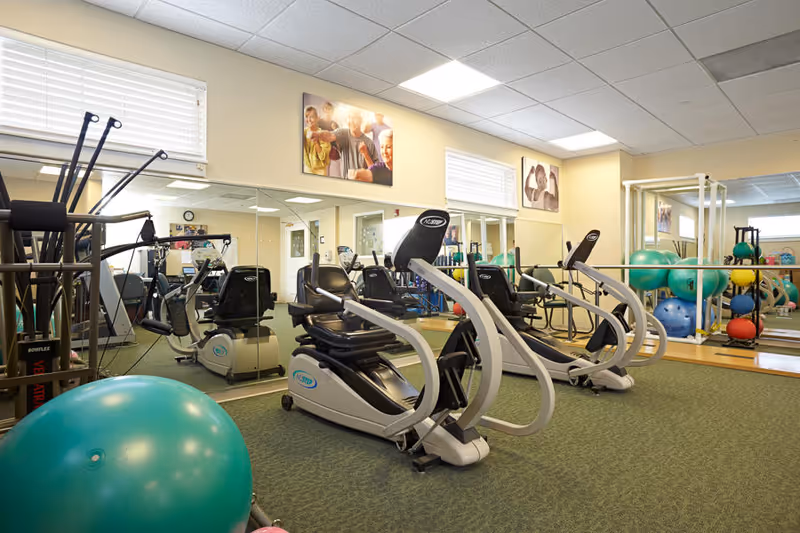 Exercise room with two recumbent stationary bikes, a large green exercise ball, various other exercise equipment, and mirrors on the walls reflecting the room. The room has beige walls, carpeted floor, and several windows near the ceiling letting in natural light. There are also framed photos of seniors on the walls.