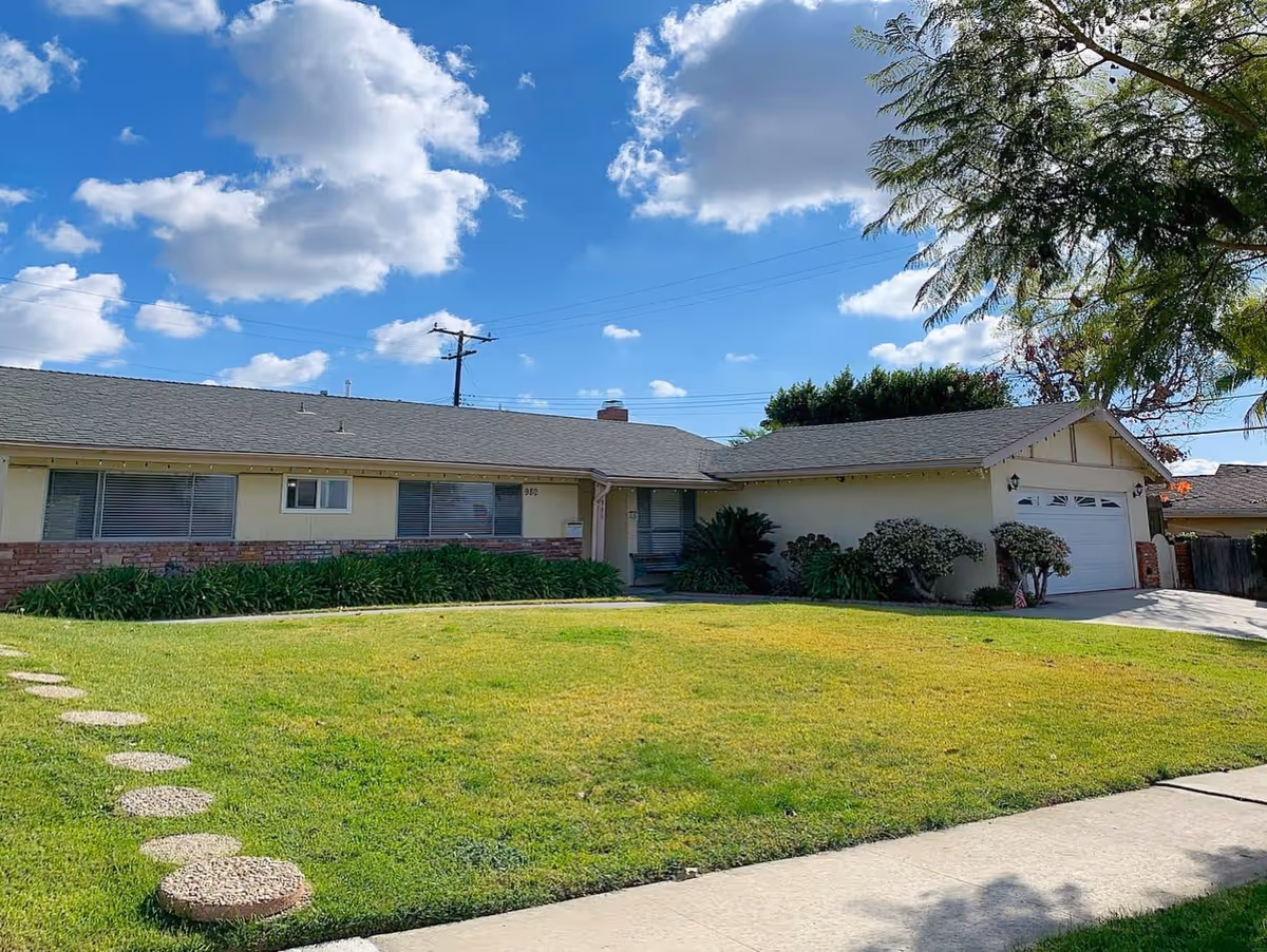 Single-story ranch-style house with a green front lawn, driveway and attached garage under a blue sky with clouds.