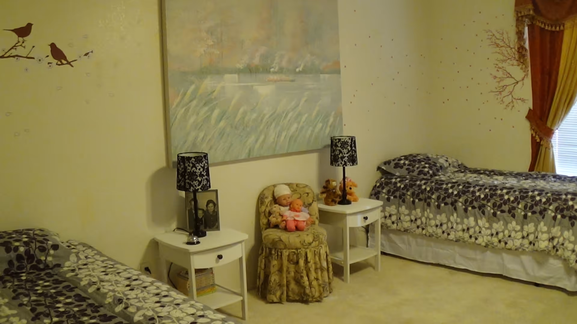 A bedroom with two twin beds covered in matching floral bedspreads. Between the beds are two white nightstands, each with a black and white patterned lamp. A small upholstered chair with a doll and a baby doll is placed between the nightstands. The walls are decorated with a large painting of a lake scene and bird decals. A window with yellow curtains is visible on the right side.