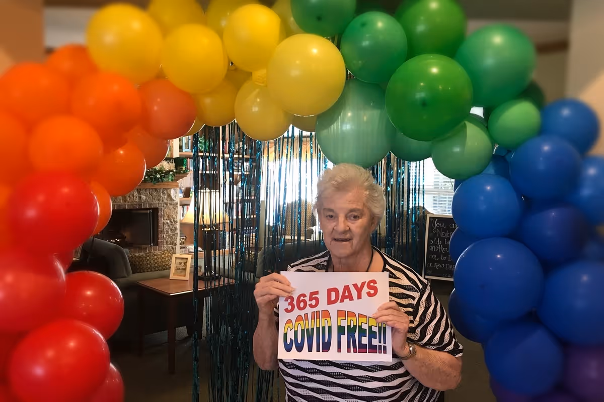 An elderly woman standing indoors under a colorful balloon arch in red, orange, yellow, green, and blue. She is holding a sign that reads '365 DAYS COVID FREE!!'. The background shows a cozy room with a fireplace, a table with a lamp, and a window with blinds.