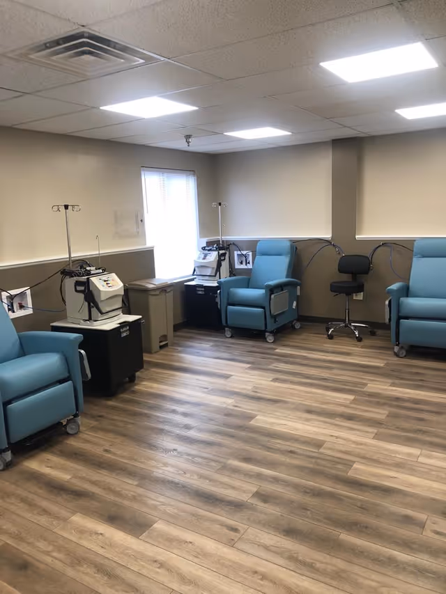 A medical treatment room with three blue reclining chairs on wheels, each positioned next to medical equipment and IV stands. The room has wood-look flooring, beige walls, and a window with blinds letting in natural light. There is also a black rolling stool and a trash bin near the window.