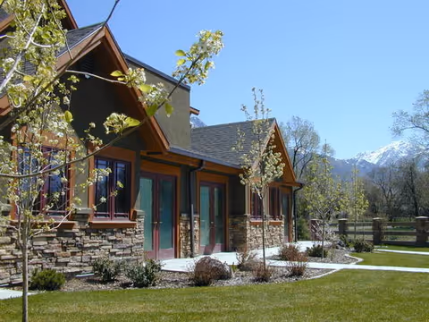 Front exterior of a low-rise building with stone accents, glass doors, landscaped lawn, young trees, and mountains in the background.