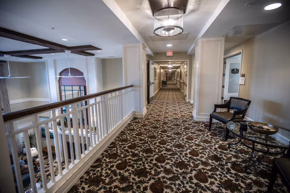 Interior hallway of a senior living facility with patterned carpet, white railing overlooking a lower level, beige walls, ceiling lights, and a small seating area with a chair and glass table on the right side.