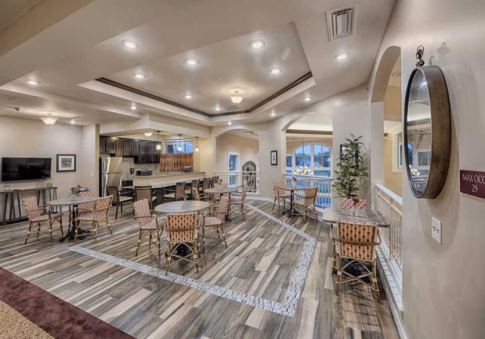 Open common dining and lounge area with multiple round tables and chairs, a kitchenette, TV, and decorative ceiling lighting.