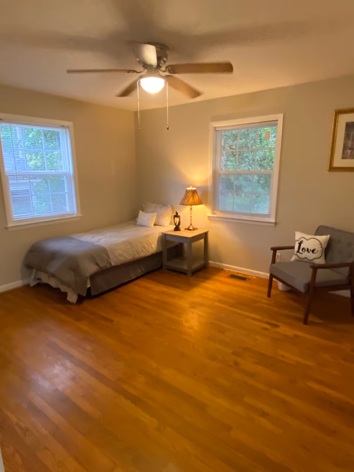 A simple bedroom with wooden flooring, a single bed with gray bedding and pillows, a small bedside table with a lamp and decorative sculpture, two windows with white blinds, a ceiling fan with light, and a gray armchair with a pillow that says 'Love'.