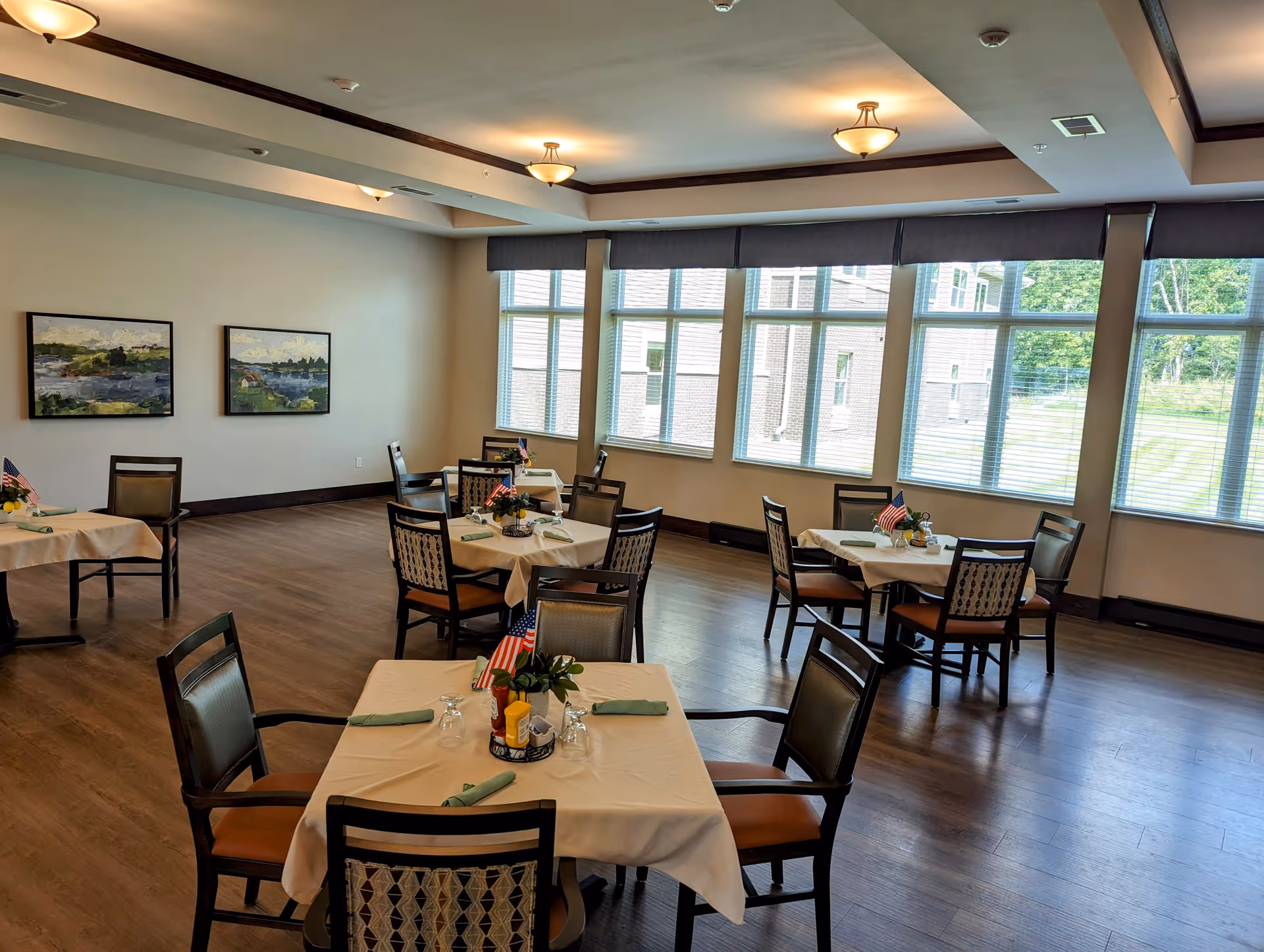 A bright dining room with several square tables covered with white tablecloths, each set with green napkins, glasses, condiments, and small flower arrangements with American flags. The room has large windows letting in natural light, wooden floors, and two landscape paintings on the wall.