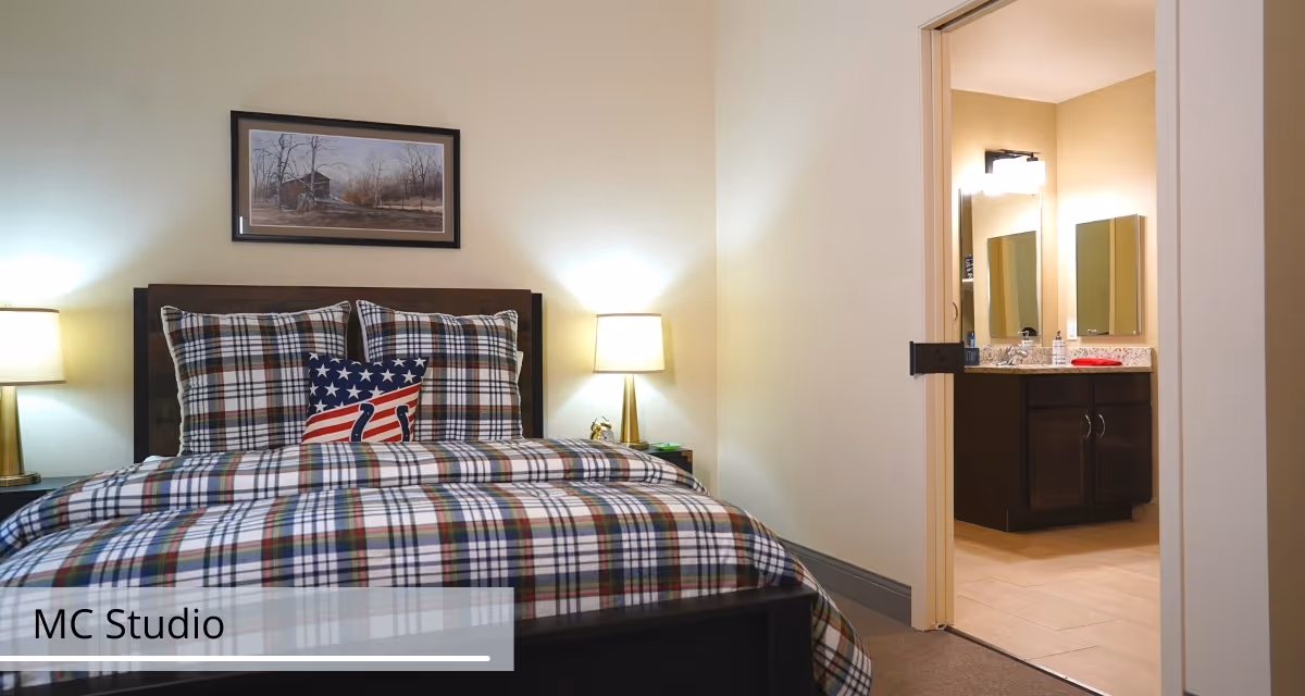 A neatly made bedroom with plaid bedding and an American-themed pillow, flanked by bedside lamps, with an open doorway revealing a bathroom vanity.