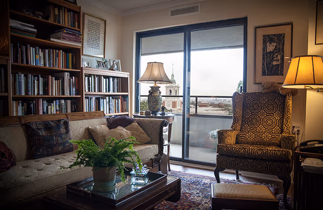 Cozy living room with a beige tufted sofa adorned with decorative pillows, a wooden coffee table with a potted fern and glass items, a patterned armchair with a matching footstool, two table lamps, a large bookshelf filled with books, and a sliding glass door leading to a balcony with a view of a church steeple and cityscape.
