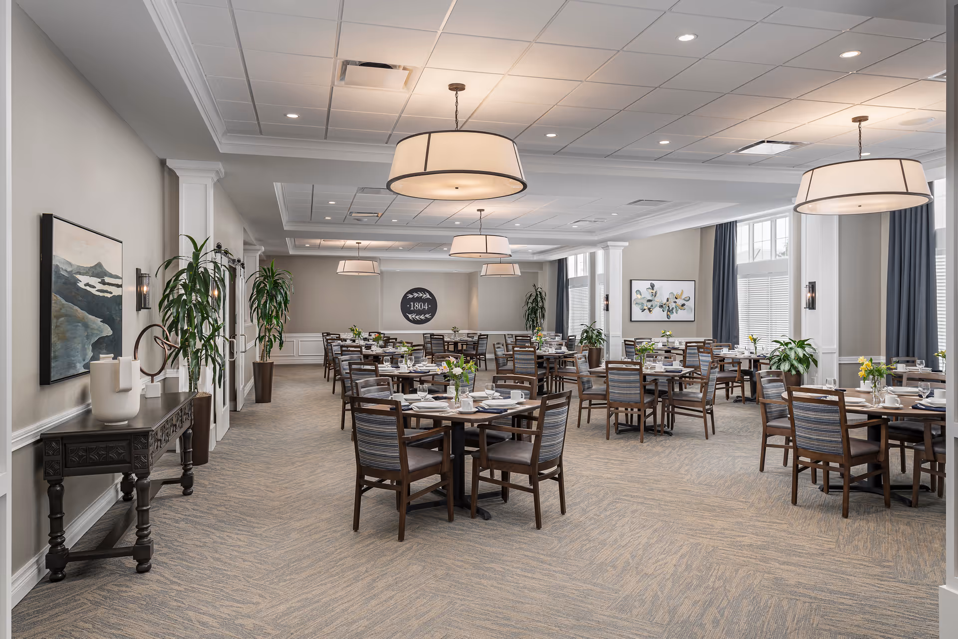 A spacious dining room with multiple round wooden tables, each set with white plates, cups, and silverware. The room features large windows with dark curtains, modern hanging light fixtures, potted plants, and framed artwork on the walls. The carpeted floor and neutral color palette create a warm and inviting atmosphere.