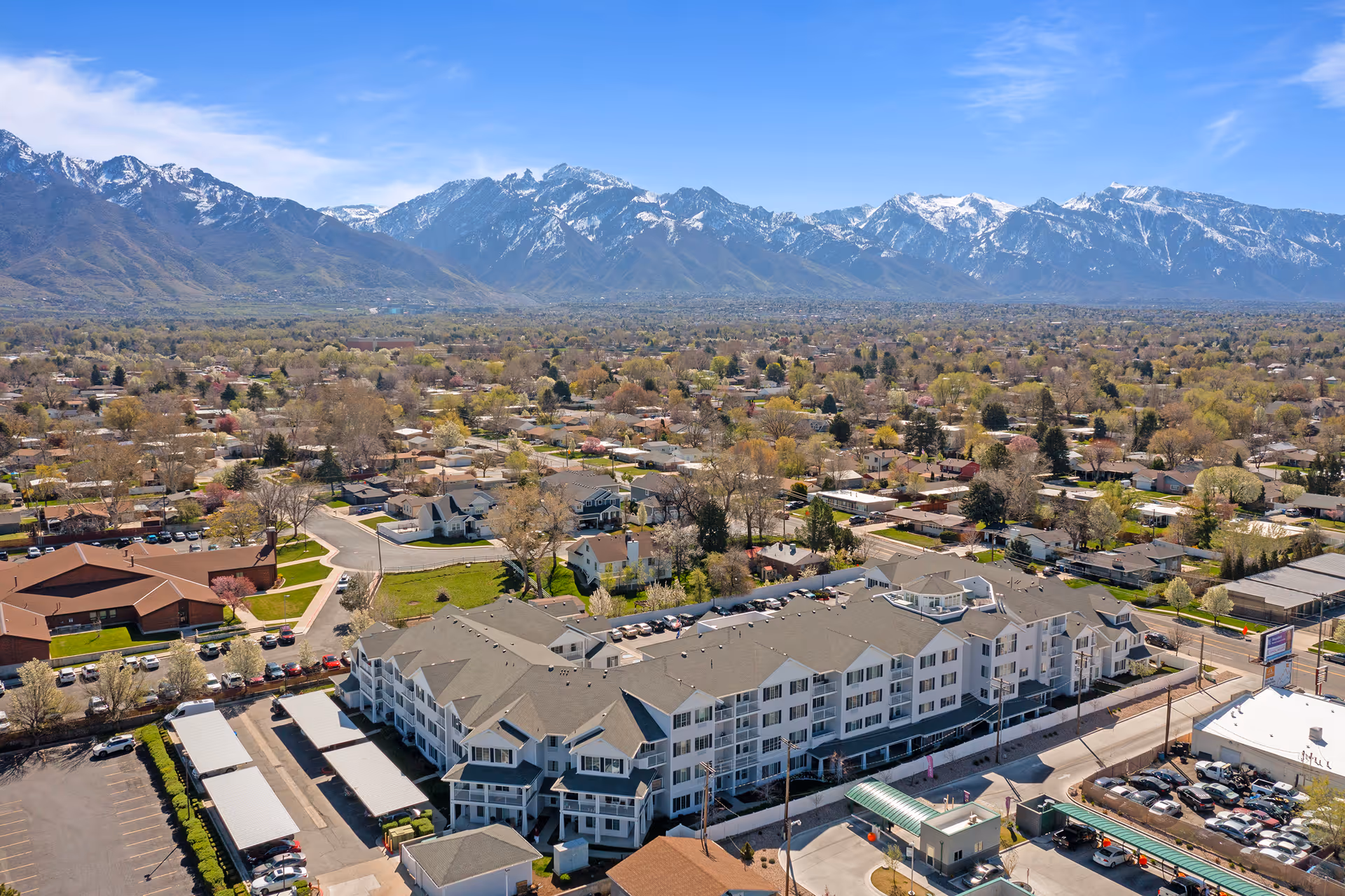 Aerial view of Olympus Ranch Gracious Retirement Living facility surrounded by residential houses with a backdrop of snow-capped mountains under a clear blue sky.