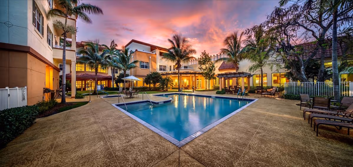 Outdoor swimming pool area at sunset surrounded by a multi-story building with palm trees, lounge chairs, tables with umbrellas, and warm lighting from the windows.