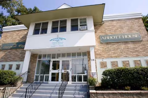 Front exterior view of Abbott Skilled Nursing and Rehabilitation Center, showing the entrance with glass doors, steps leading up to the doors, and signs on the building reading 'Abbott House' and 'Abbott Skilled Nursing and Rehabilitation Center'.
