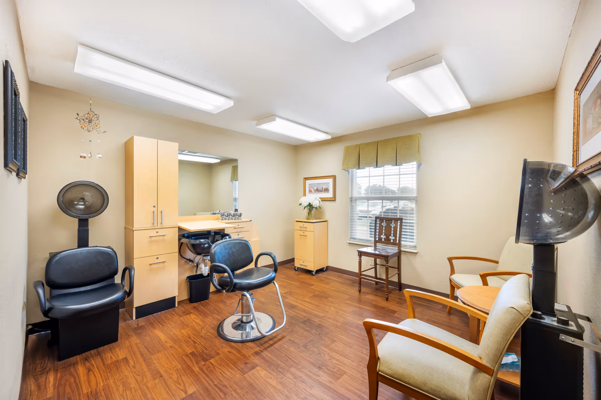 Interior of a hair salon room in a senior living facility with two black salon chairs, a hair washing station, wooden cabinets, a large mirror, two hair dryers, and seating area with beige chairs and a small round table near a window with blinds and a green valance.