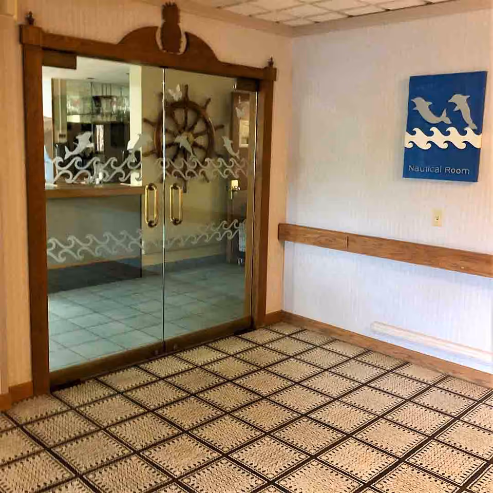 Interior view of a hallway with a nautical theme, featuring glass double doors with wave and dolphin designs, a wooden ship wheel decoration behind the doors, a patterned carpet, and a blue sign on the wall labeled 'Nautical Room' with dolphin and wave graphics.
