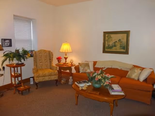 Cozy living room with an orange sofa, patterned armchair, wooden coffee table with a floral centerpiece, and a lit table lamp.