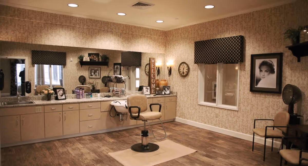 A well-lit salon-style interior with a central styling chair, long vanity with sinks and mirrors, framed photos, and patterned wallpaper.