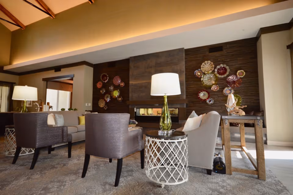 A cozy living room area with modern furniture including a gray sofa, two gray armchairs, and a beige armchair around a round side table with a green glass lamp. The room features a contemporary fireplace with decorative colorful glass wall art on a dark wooden panel behind it. The space is warmly lit with natural light coming from the right side.