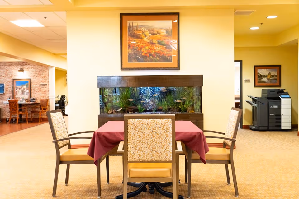 A seating area with a square table covered by a maroon tablecloth and four chairs with patterned upholstery. Behind the table is a large aquarium filled with fish and aquatic plants, set against a cream-colored wall. Above the aquarium hangs a framed painting of a scenic landscape with trees and a field. To the left, there is another seating area with a wooden table and chairs, and to the right, a large office printer is visible next to a framed picture on the wall.