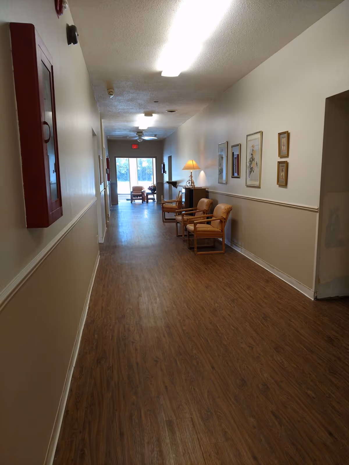A long hallway in a senior living facility with wood-patterned flooring and beige walls. On the right side, there are several framed pictures, a table with a lamp, and two cushioned chairs. At the end of the hallway, there is a glass door letting in natural light and a small seating area with chairs and a table.