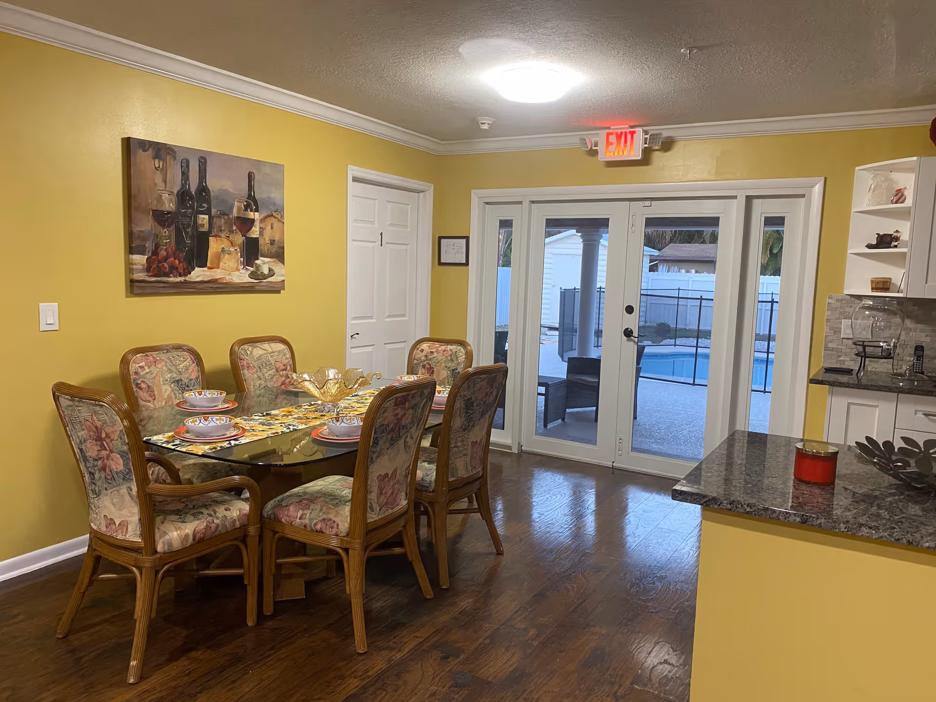 Dining area with six floral-upholstered chairs around a glass table, yellow walls, and glass doors leading to a pool patio.