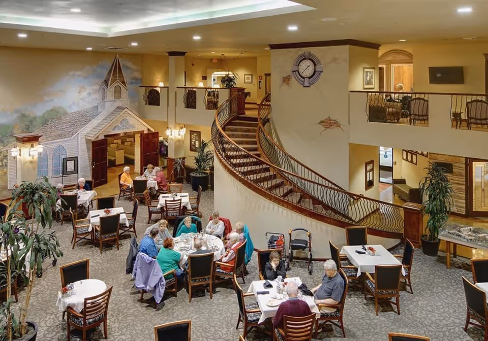 A spacious dining area in a senior living facility with several elderly people seated at tables, eating and conversing. The room features a large decorative mural of a church on one wall, a curved staircase with ornate railing, and a clock mounted on a beige wall. There are plants and multiple tables with chairs arranged throughout the carpeted room.
