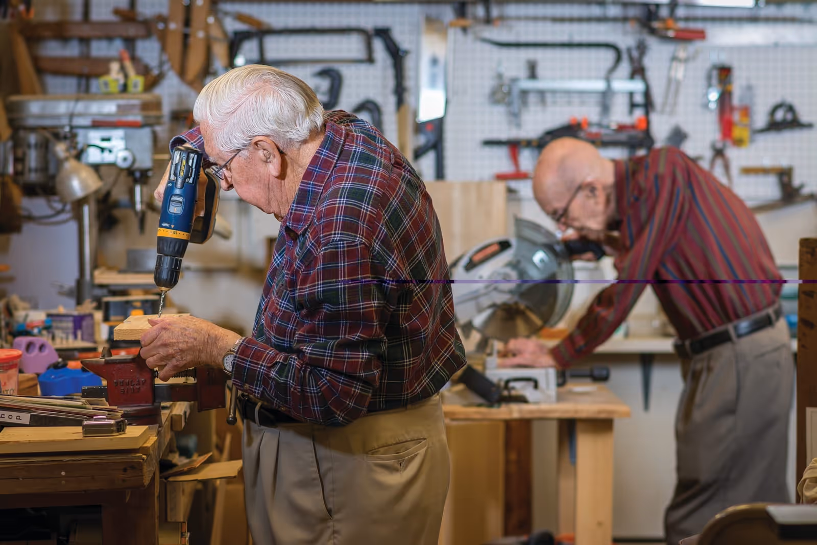 Two elderly men working in a woodworking shop. One man in the foreground is using a power drill on a piece of wood held in a vise, while the other man in the background is operating a miter saw. The workshop is equipped with various tools hanging on a pegboard and workbenches.