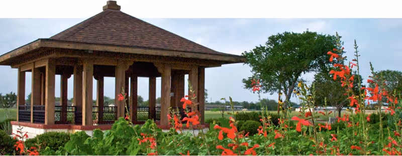 A wooden gazebo with a shingled roof situated in a garden area with green bushes and red flowers, with trees and open fields in the background under a cloudy sky.