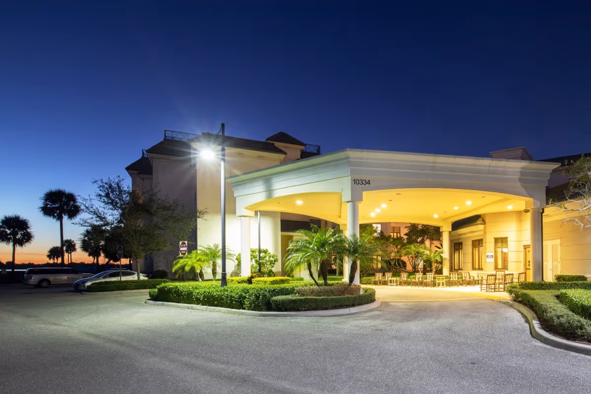 Illuminated entrance canopy and driveway of a senior living building at dusk with palm trees and seating.