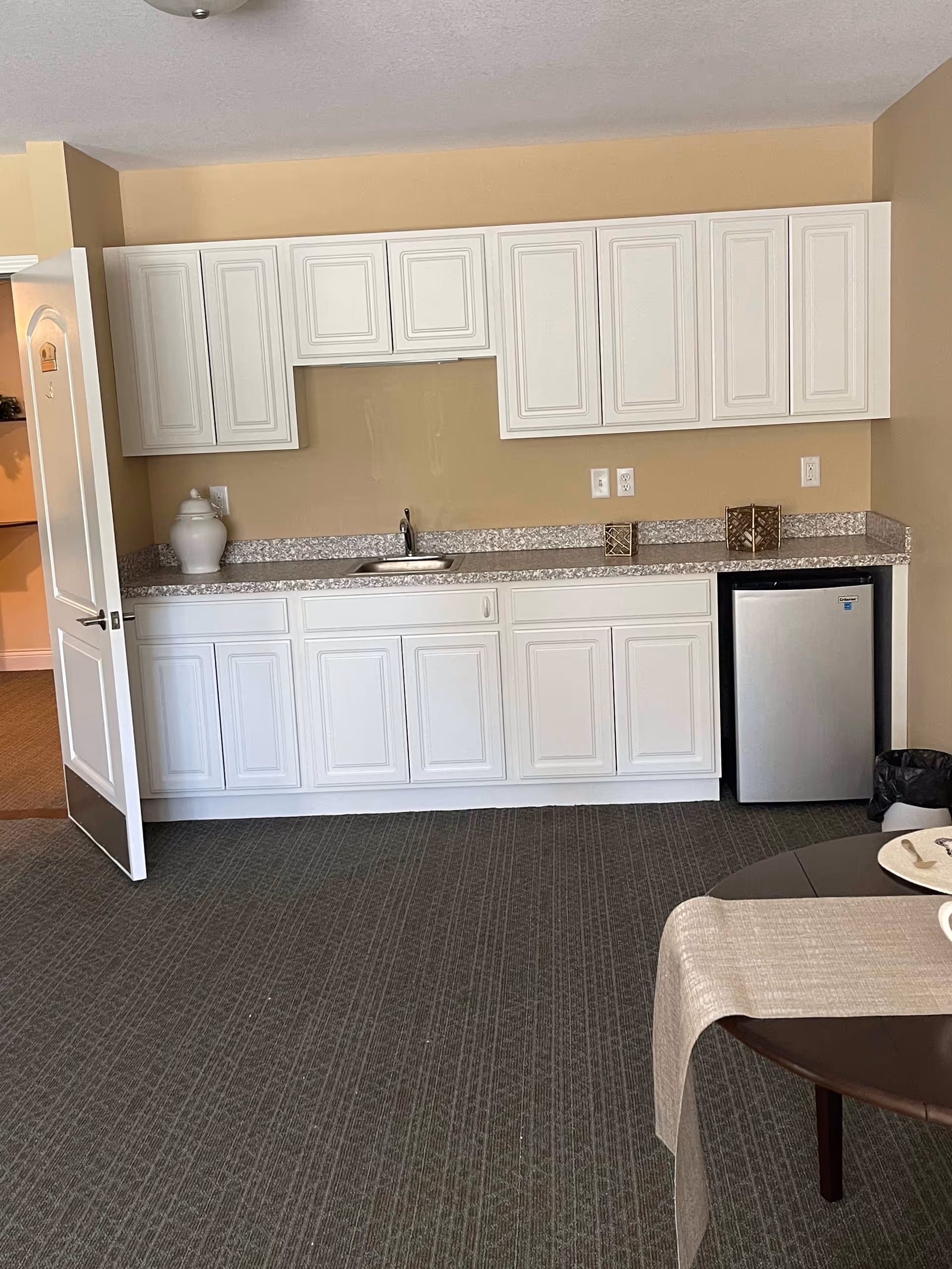 Interior view of a kitchenette area with white cabinets, a small sink, a mini refrigerator, and a countertop with decorative items. The room has beige walls and a carpeted floor. A round table with a beige table runner and plates is partially visible in the foreground.