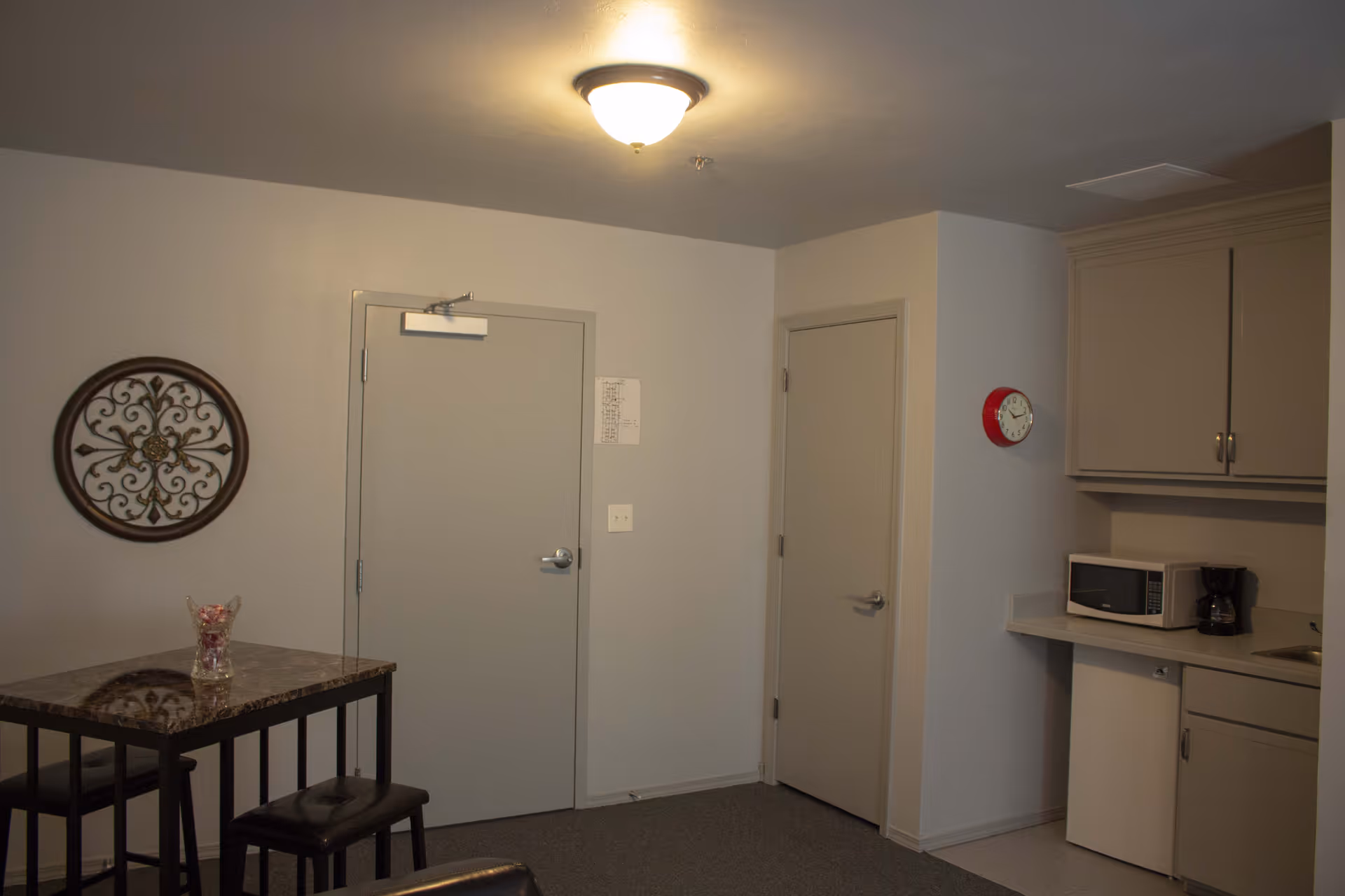 Small interior kitchenette and dining area with a high table and stools, two doors, wall decor, and a microwave and coffee maker on the counter.