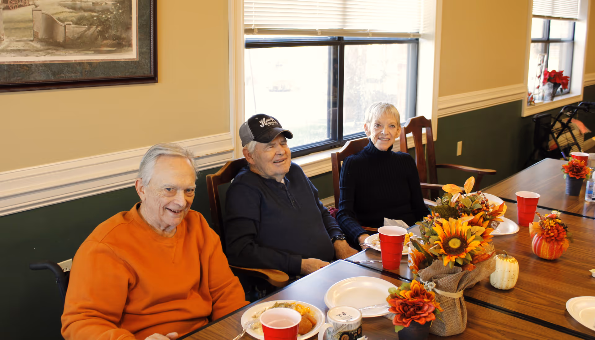 Three older adults seated at a decorated dining table with plates, red cups, and fall floral centerpieces in a bright room.