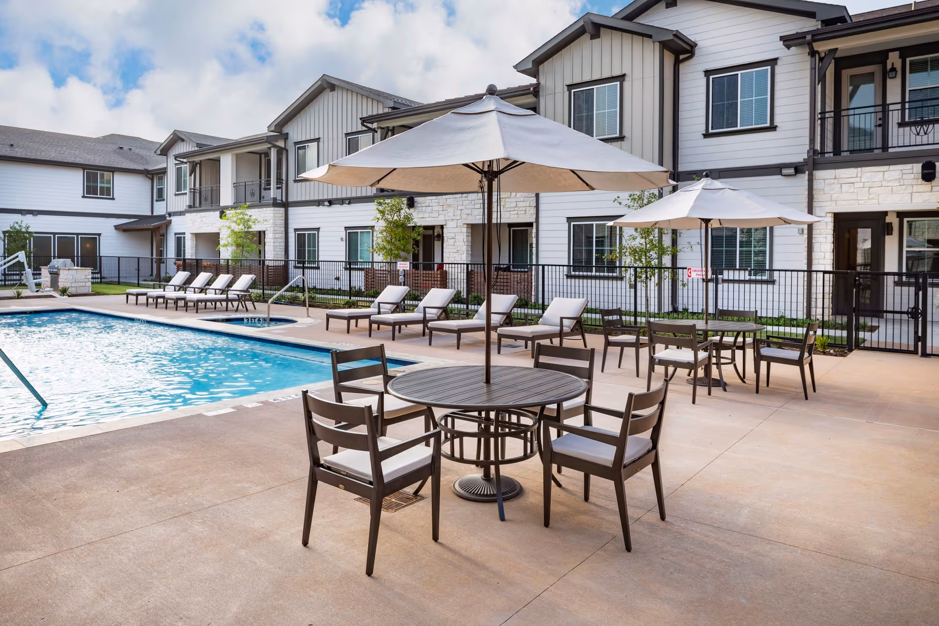 Outdoor pool patio with round tables, umbrellas and lounge chairs in front of a two-story senior living building.