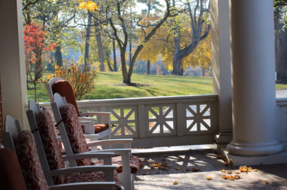 A porch area with white rocking chairs that have red patterned cushions, overlooking a grassy area with trees showing autumn foliage in shades of yellow and red.