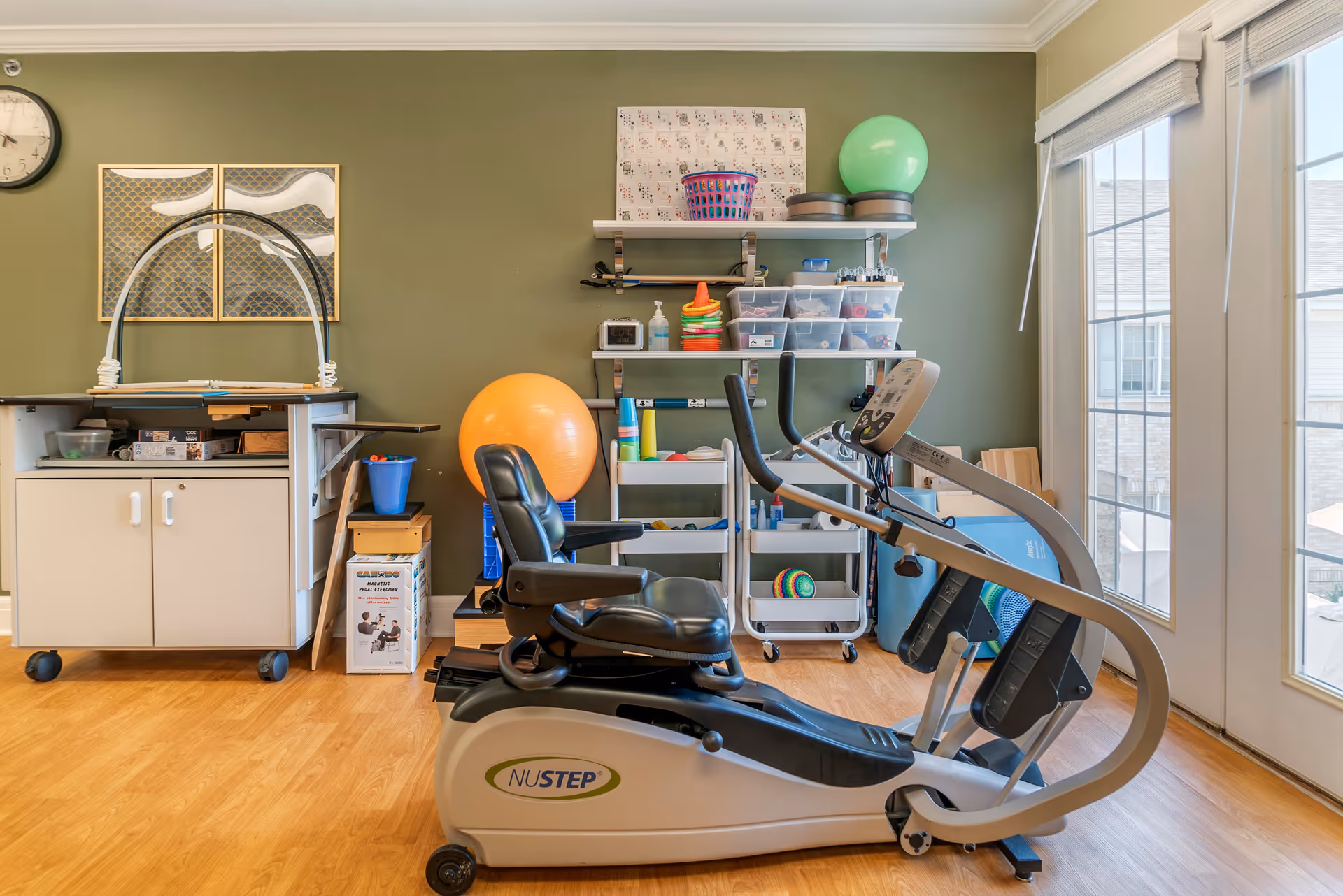 A physical therapy or exercise room with a NuStep recumbent cross trainer in the foreground. Behind it are shelves with various exercise and therapy equipment including an orange exercise ball, green exercise ball, cones, and plastic bins. The room has wooden flooring, a green wall with a clock and artwork, and large windows letting in natural light.